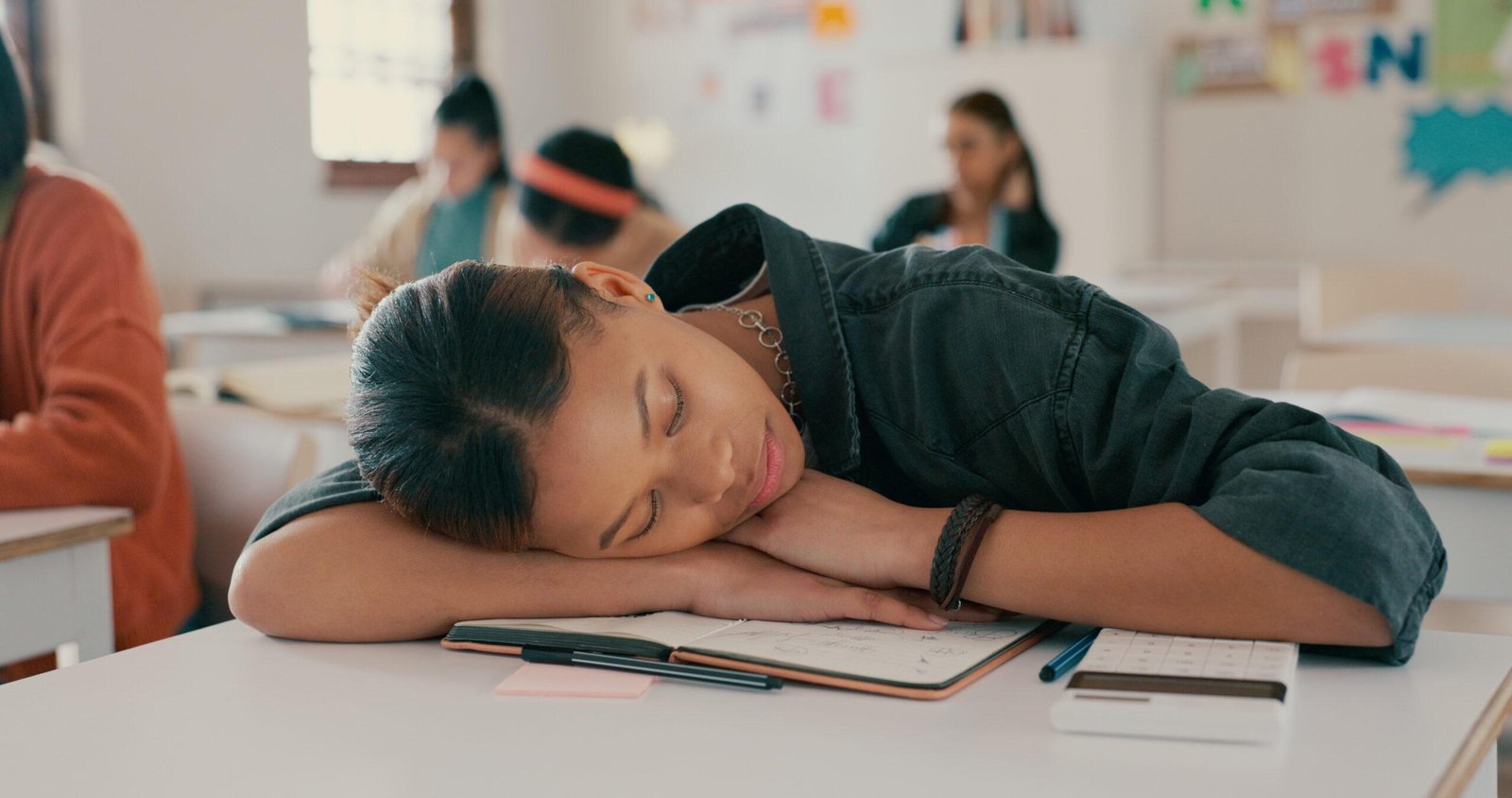 Teenage student resting her head on a desk in a classroom, surrounded by classmates engaged in studying.